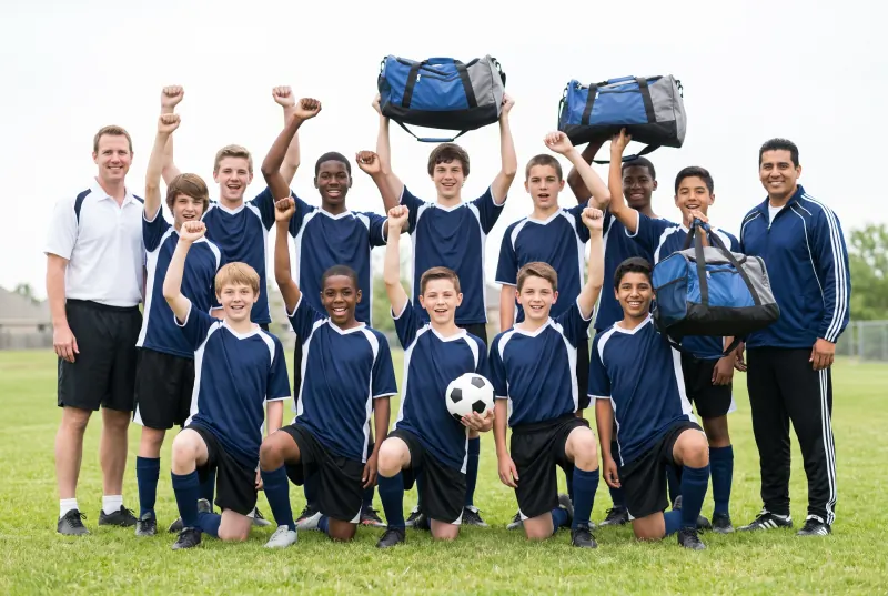 A youth soccer team and two coaches celebrating on a field, holding up new equipment bags